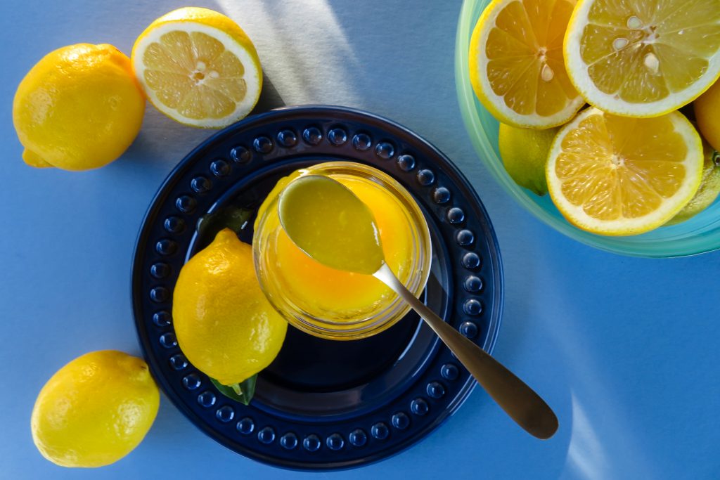 Bright blue background with filters sun in the upper left-hand corner leading to the bright yellow lemon curd on a navy blue plate surrounded by whole and cut lemons.
