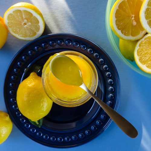 Bright blue background with filters sun in the upper left-hand corner leading to the bright yellow lemon curd on a navy blue plate surrounded by whole and cut lemons.